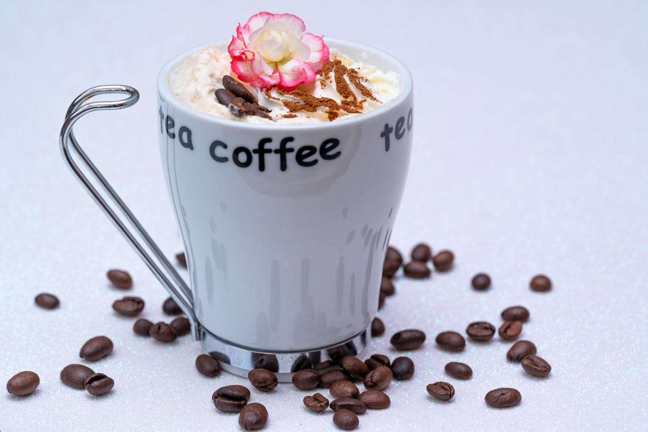 A stylish coffee mug with whipped cream, cinnamon, a pink flower, and scattered coffee beans on a white background.